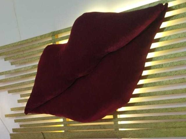 SahraValley, vestita con un abito rosso scuro e vellutato, posa davanti a uno sfondo di listelli di legno illuminati da un giallo brillante. luci. Le pieghe del vestito creano una silhouette sensuale e fluida..