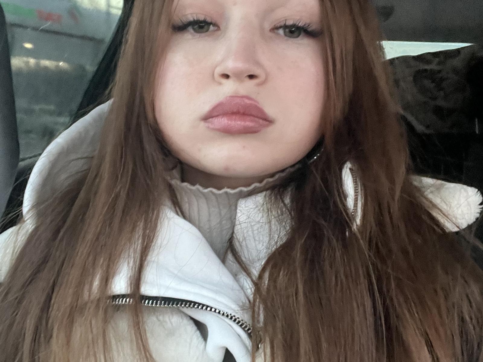 Close-up of IzolaCrabtree, a fair-skinned woman with long brown hair, wearing a white coat, looking directly at the camera with slightly parted pink lips, creating a subtle sexual tension.