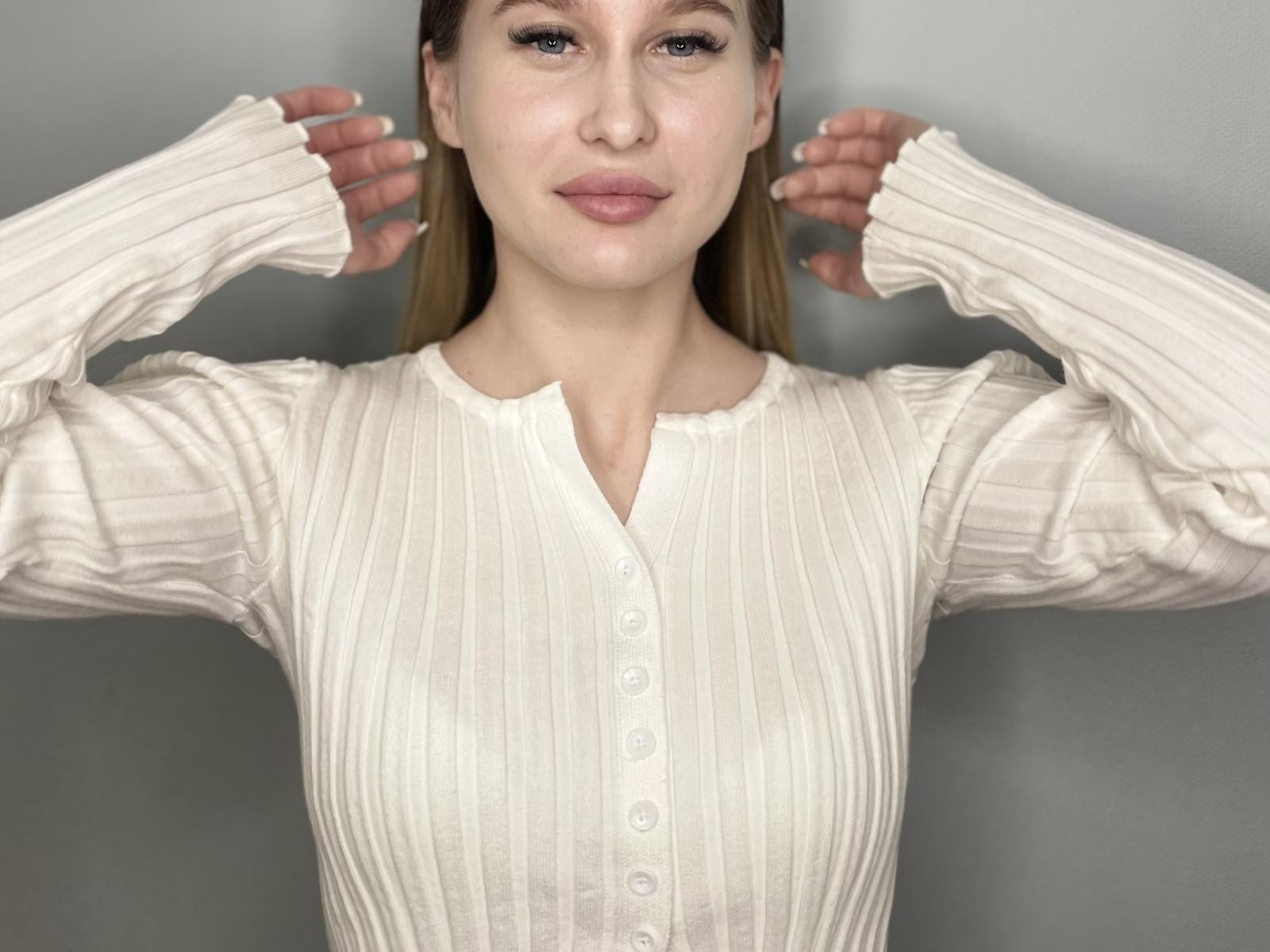 Digital photo of DeenaSerpe, light-skinned woman with brown hair, wearing a white, ribbed, button-up shirt, hands raised to her shoulders, blue eyes, slight smile, soft lighting, gray background.