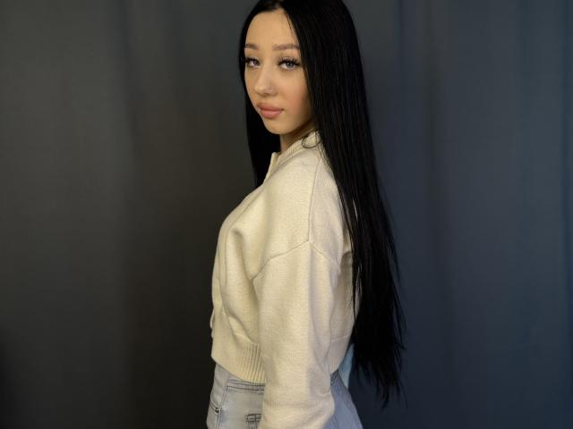 Fotografía de FredLenzen, una joven con cabello largo y negro, que lleva un suéter blanco y pantalones grises, y que está mirando hacia arriba. con un gesto sensual. Fondo negro. Tensión sexual. Increíblemente atractivo..