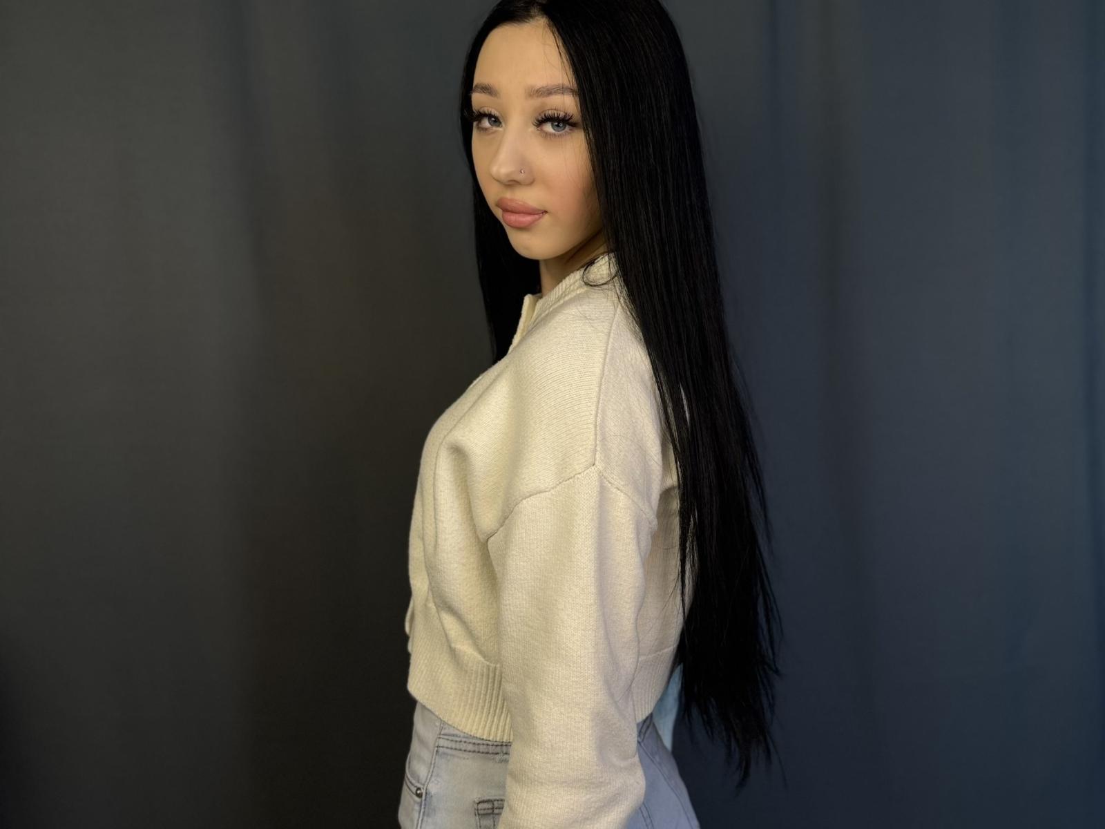 Fotografía de FredLenzen, una joven con cabello largo y negro, que lleva un suéter blanco y pantalones grises, y que está mirando hacia arriba. con un gesto sensual. Fondo negro. Tensión sexual. Increíblemente atractivo..