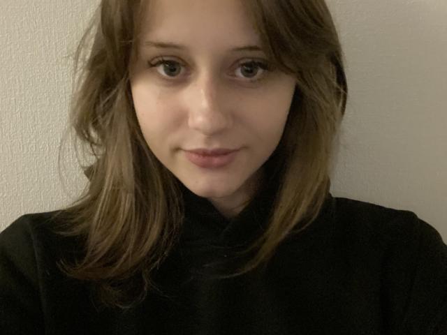 Close-up van OkKeilty met bleke huid, bruine ogen en bruin haar, die een zwart shirt draagt. Ze kijkt recht in de camera met een lichte, zelfverzekerde glimlach. Duidelijke seksuele spanning..