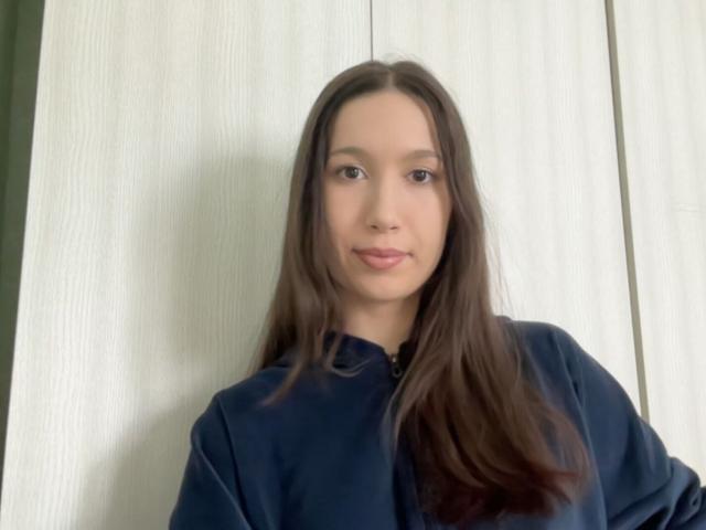 Photograph of DavidBacco, a young woman with long brown hair, wearing a black shirt, standing against a white, textured background. She gazes directly at the camera with a subtle smile.