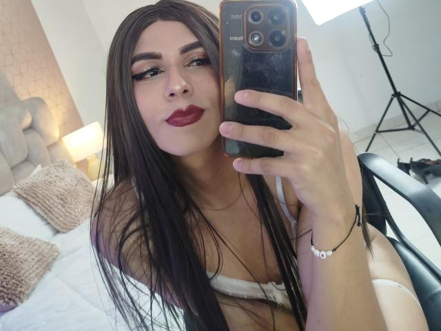 SahraValley macht ein Selfie vor einem Spiegel mit ihrem Smartphone und trägt dabei weiße Dessous, die ihren Ausschnitt zeigen. Sie hat langes, schwarzes Haar, dunkelroten Lippenstift und hält das Telefon in ihrer rechten Hand. Ihr Ausdruck ist verführerisch..