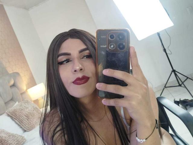 SahraValley, une femme séduisante avec de longs cheveux noirs et du rouge à lèvres, prend un selfie avec un téléphone Samsung, en le tenant en l'air. devant une lumière de studio très lumineuse. Sa poitrine est visible..