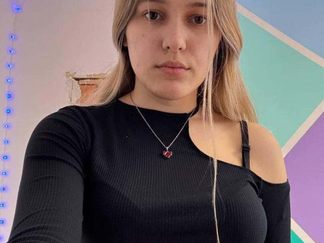 BelleDubicki, avec ses cheveux bruns clairs, portant un haut noir côtelé avec une bretelle tombant d'une épaule, regardant directement... caméra. Elle porte un collier avec un pendentif, on voit sa poitrine, et elle a une expression sérieuse..