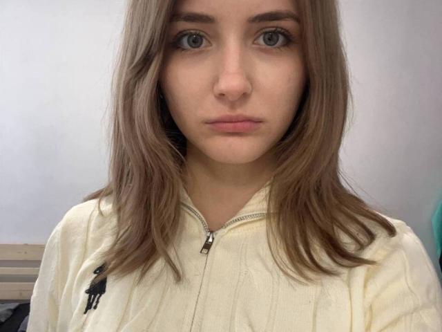 Close-up of DelorisBueno, a young woman with fair skin, blue eyes, and brown hair, wearing a cream-colored zip-up shirt. Her expression is neutral, with a subtle hint of sexual tension.