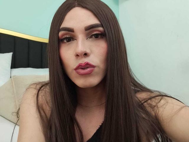 Gros plan de SahraValley, une femme brune avec de longs cheveux lisses, des lèvres pulpeuses et un maquillage prononcé, qui fait une moue sexy. une personne, portant un haut noir, sur un fond blanc et noir.