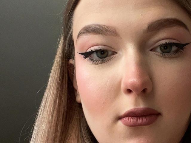 Close-up of NadaHenig's face, centered on her eyes and nose. She has light skin, green eyes with black winged eyeliner, and full, pinkish lips. Her straight, blonde hair falls to the left. Her eyebrows are neatly shaped and slightly arched. The lighting is even, highlighting her smooth skin texture. The background is dark and out of focus. NadaHenig's expression is neutral, with a slight downward tilt of her lips.