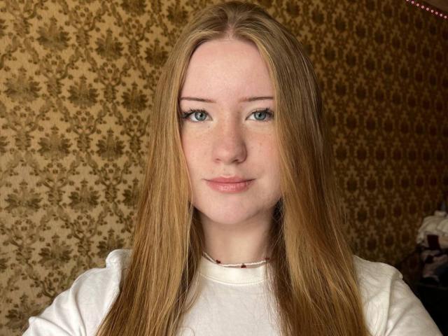 TashinaDufort has long, straight, blonde hair, blue eyes, and a slight smile. She's wearing a white shirt with a small red bead necklace. Her ass is not visible.
