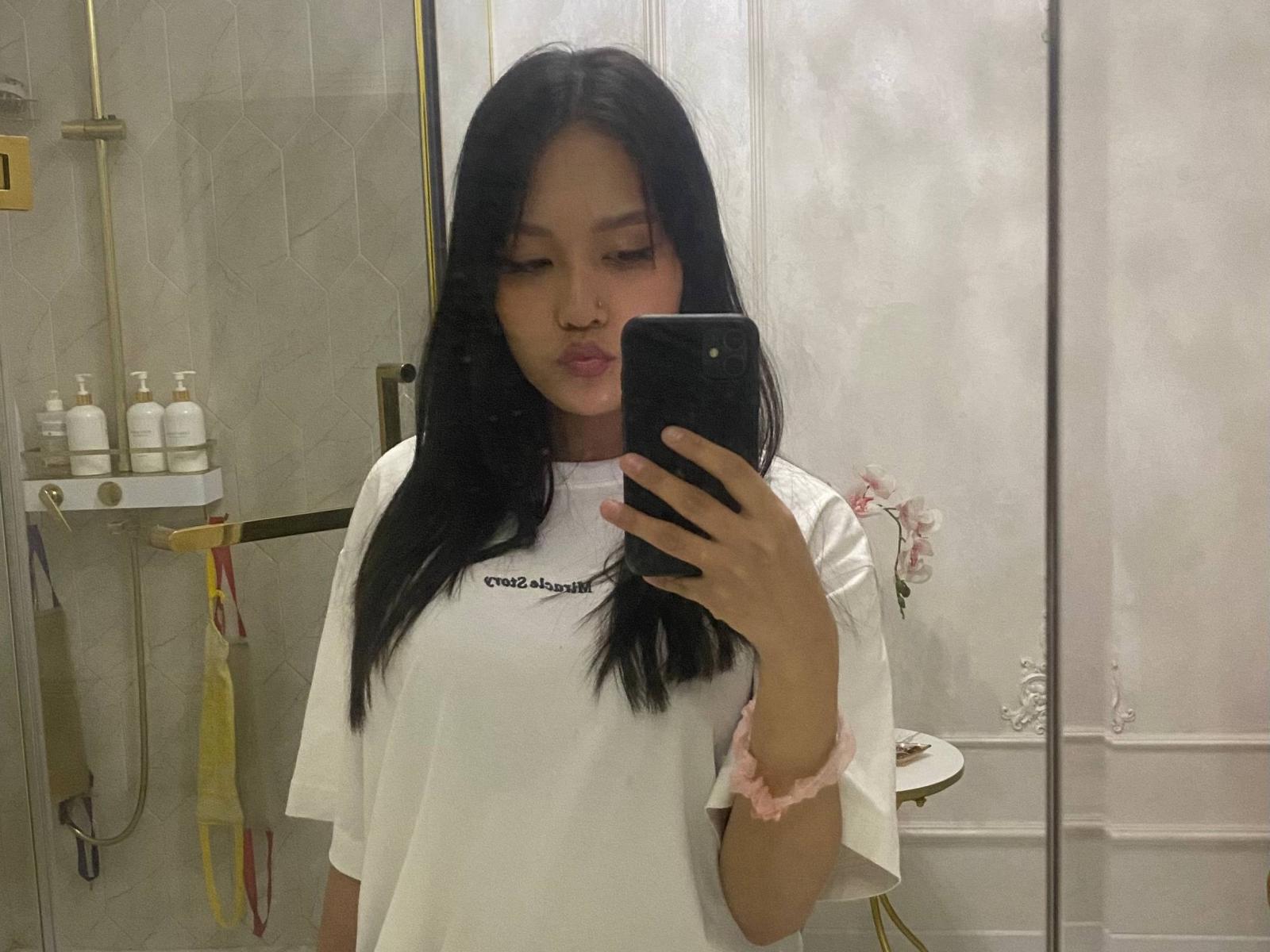 Teressa Calizo tira uma selfie em um banheiro, segurando um telefone preto com a mão direita. Ela está vestindo uma camiseta branca, tem cabelo preto e um elástico rosa no cabelo. Ela está fazendo um leve biquinho. Prateleiras com produtos de higiene pessoal ao fundo..