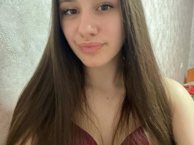 Close-up van WavaBlauman, een jonge vrouw met lang bruin haar, lichte huid en kleine borsten, die een donkerrode, strapless top draagt. bovenkant, lichte glimlach, zachte verlichting, zichtbare textuur op de muur.