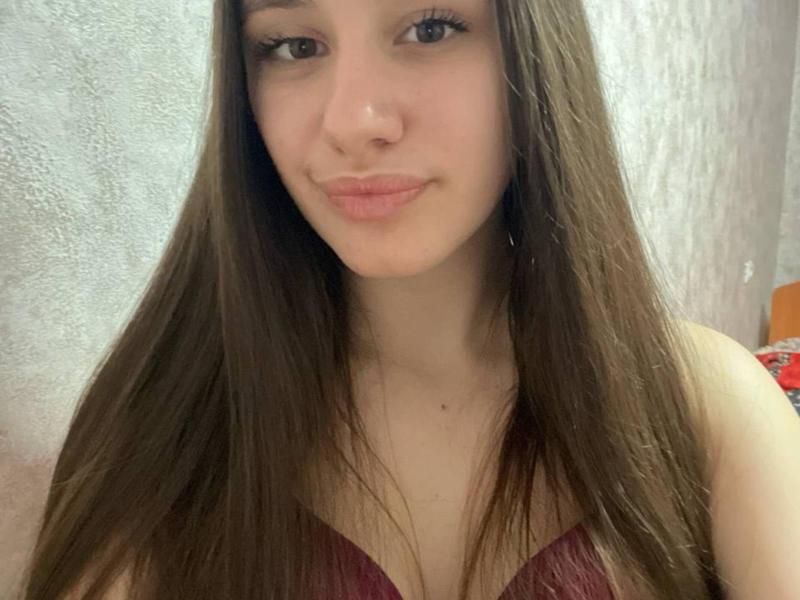 Gros plan de WavaBlauman, une jeune femme aux longs cheveux bruns, à la peau claire et à la petite poitrine, portant un bustier bordeaux sans bretelles. en haut, léger sourire, éclairage doux, texture visible sur le mur.