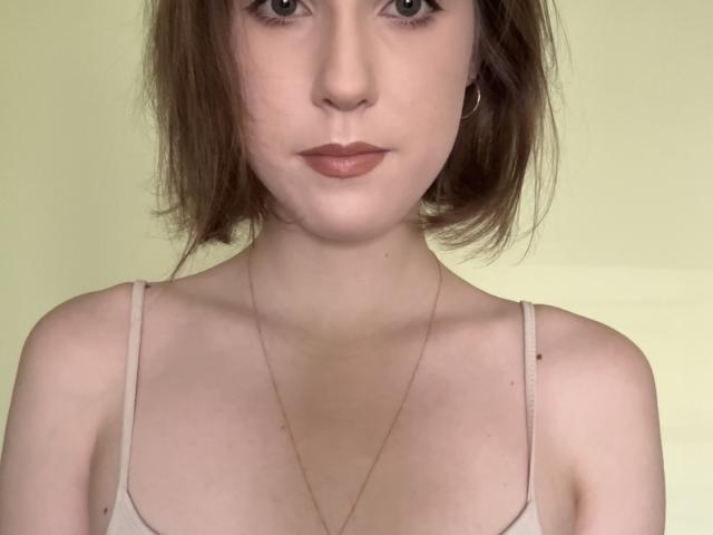 Close-up of KenaCly, a young woman with pale skin and brown bob haircut, wearing a white tank top, thin gold necklace, and hoop earrings, with a serious expression and direct gaze.