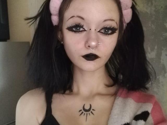 NicolaRichburg with black hair, pink puffy headband, black eyeliner, black lipstick, nose ring, black tank top, sun tattoo on chest, and a serious expression. Boobs visible, small chest.
