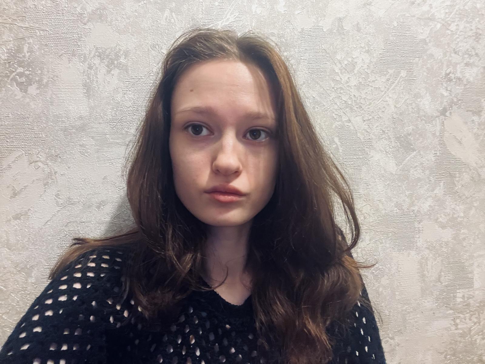 JessieYen has long, brown hair, pale skin, and a neutral expression. She wears a black, patterned top against a textured, white background. Her wide eyes and slightly parted lips add tension.