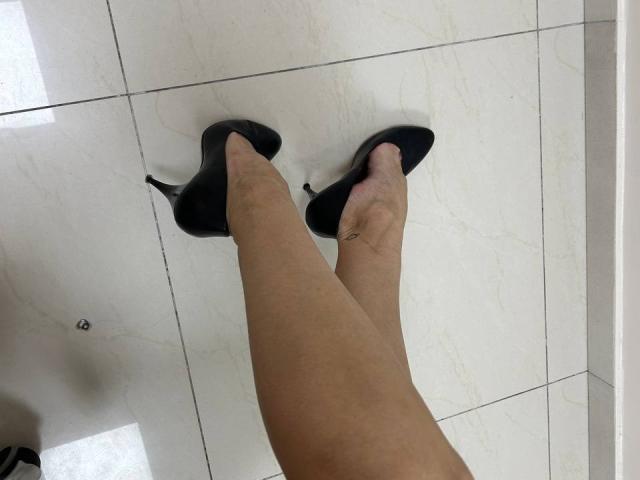 Fotografía de primer plano de las piernas de MoonLiza, con piel clara, usando zapatos de tacón alto negros, de pie sobre un suelo de baldosas blancas brillantes. suelo.
su tobillo derecho lleva un anillo de plata. Piernas increíblemente atractivas..