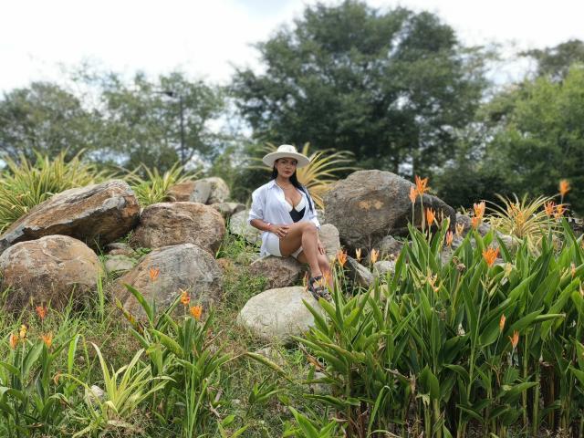 JessicaFiore assise parmi de gros rochers et des fleurs orange, portant une chemise et un chapeau blancs, les jambes croisées, exposant ses entrejambe, arrière-plan forestier, lumière vive, environnement naturel, tension sexuelle..