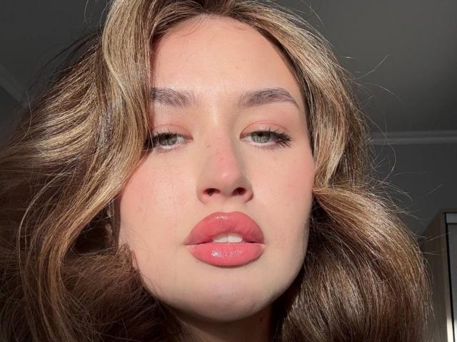 Close-up van EstherDambrose, met een lichte huid, glanzende roze lippen, halfgesloten ogen en golvend bruin haar. Het zonlicht accentueert haar gezicht, waardoor ze een licht verleidelijke, intense uitdrukking krijgt. Geconcentreerd op haar gezicht..