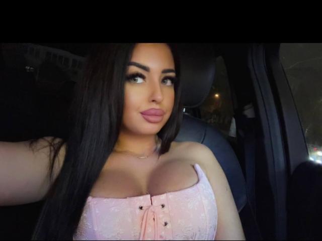 Fotografía de OlyviaDelight, sexy y voluptuosa, con cabello largo y negro, vistiendo un corsé rosa ajustado que resalta su figura. senos grandes. Ella tiene labios carnosos, mucho maquillaje y una expresión sensual, inclinándose hacia adelante..