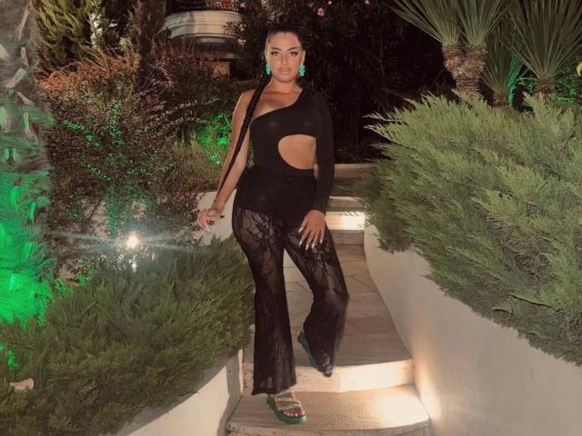 OlyviaDelight stands on a lit stone path, wearing a black, cut-out top and sheer pants, with green earrings. Her hand rests on her hip, exuding confident, sexy vibes.