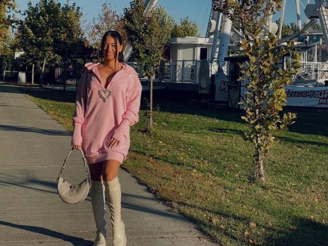 OlyviaDelight está de pie en una acera a pleno sol, vistiendo una sudadera rosa, botas blancas hasta la rodilla y sosteniendo una bolso de mano. Tiene una figura curvilínea y una expresión segura. Proviene de un entorno suburbano..