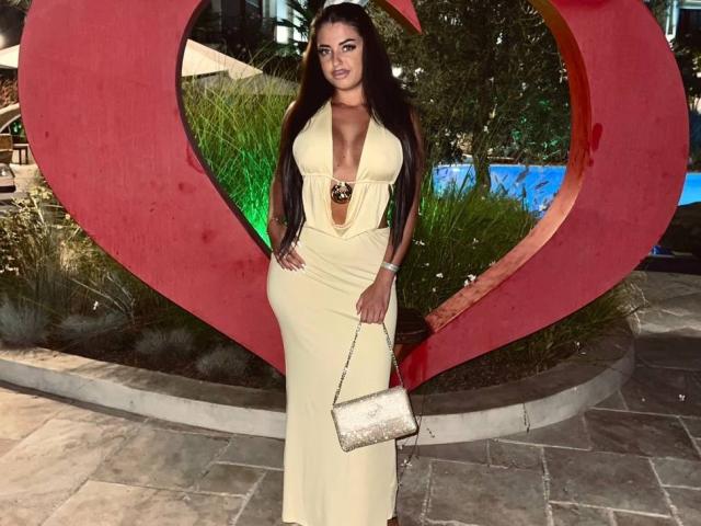 OlyviaDelight stands in front of a large red heart sculpture, wearing a tight, low-cut, cream-colored dress that accentuates her curvy figure and large breasts. She holds a white clutch, looking sexy.