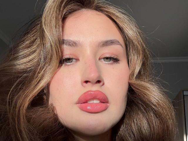 Primo piano del volto di EstherDambrose, con capelli castano lucidi, labbra piene rosate leggermente dischiuse e occhi seducenti. La sua pelle è chiara e si vedono i pori, e ha un'espressione seducente. La luce intensa mette in risalto i suoi lineamenti..