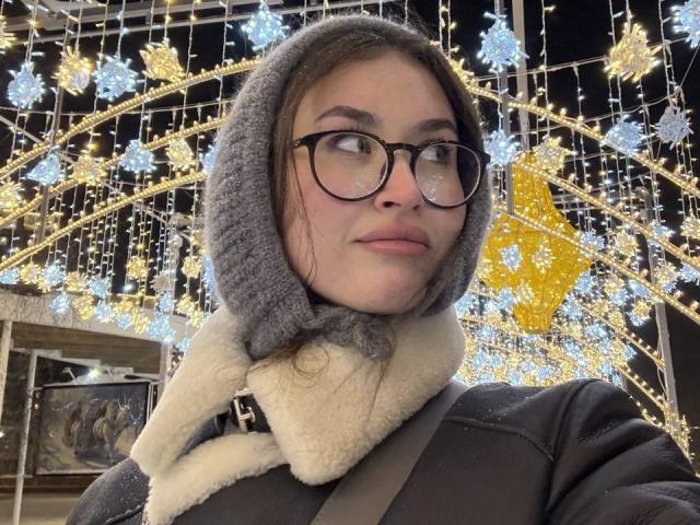 Photographie d'Esther Dambrose, une femme à la peau claire et portant des lunettes, vêtue d'un manteau gris à capuche avec un bordure en fourrure blanche. debout devant une décoration lumineuse extérieure festive avec des flocons de neige jaunes et blancs.