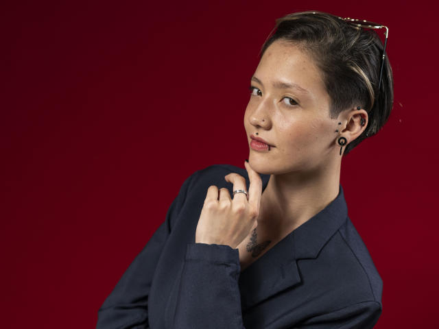 Fotografía de LissyLuxe con cabello negro corto y peinado hacia atrás, vistiendo un blazer azul marino oscuro, posando con la mano cerca de la barbilla, de color rojo. fondo, mirada intensa, sonrisa sutil, ligero sudor en la cara, vibra de confianza..