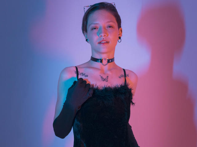 LissyLuxe mit kurzen, braunen Haaren, trägt ein schwarzes Kleid aus Federn, eine schwarze Halskette mit Herzanhänger und schwarze Handschuhe. steht vor blau-rosa Beleuchtung, im rechten Bereich im Schatten. Verdammt heiß..