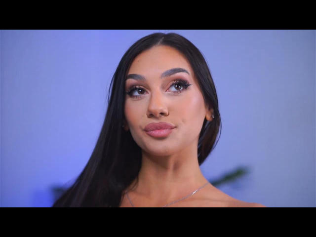 Foto van NinaSolenne, een aantrekkelijke vrouw met lang zwart haar, volle lippen en dikke wenkbrauwen. Ze kijkt omhoog met een lichte glimlach, blauwe achtergrond, lichte onscherpte aan haar rechterkant..