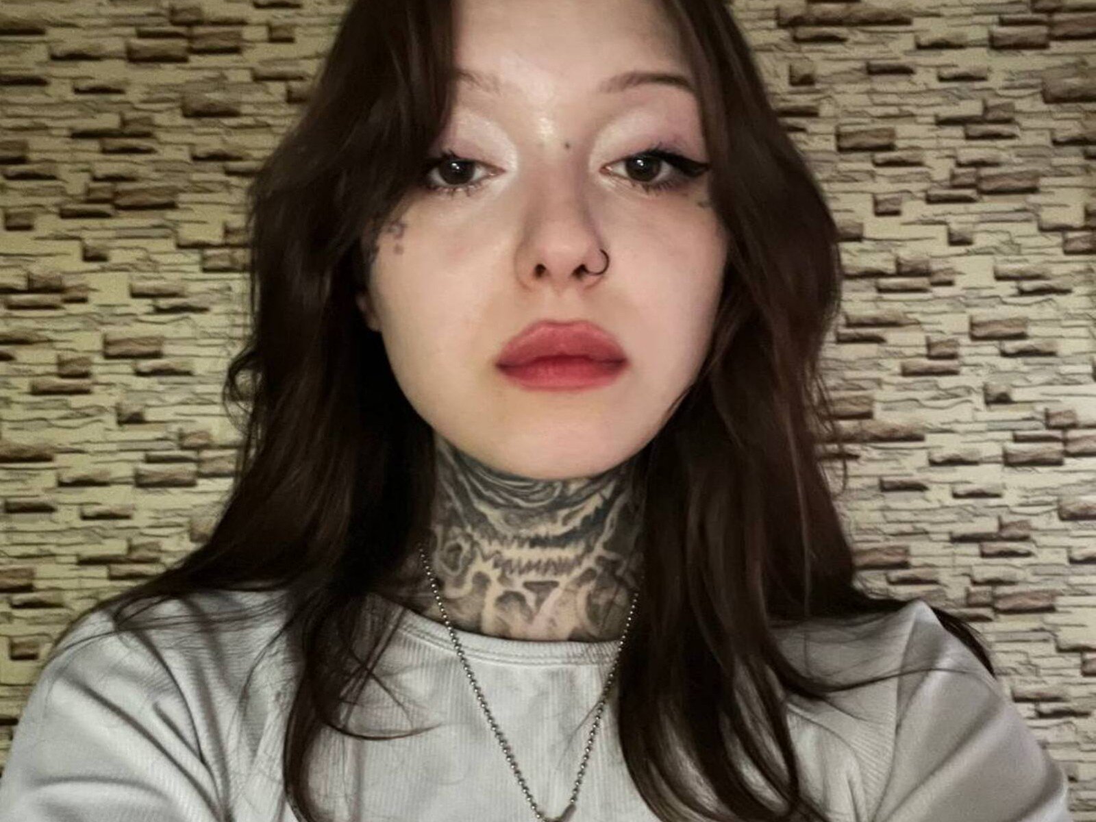 Close-up of GermaineMagraw with pale skin, dark brown hair, red lips, nose ring, and extensive neck tattoo. Wearing white shirt, silver necklace. Textured beige background. Fucking sexy look.