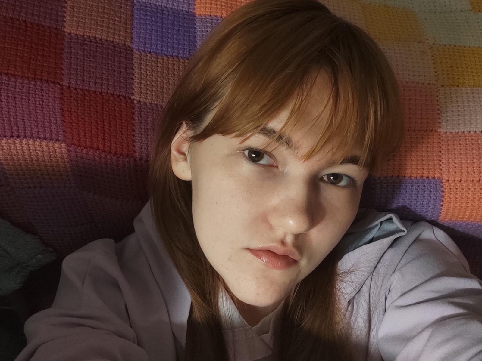 Close-up van SheritaChieng, met een lichte huid en bruin haar, liggend op een kleurrijke, geruite bank. Ze draagt een lichtgrijze hoodie en kijkt recht in de camera met een neutrale uitdrukking. Zachte schaduwen accentueren haar gezicht.