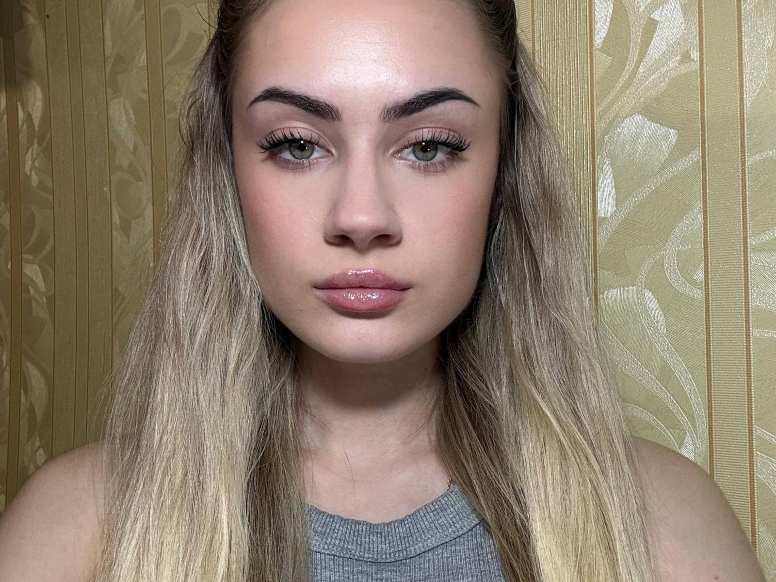 Fotografía de primer plano de Albertina Vacha, con cabello rubio largo, piel clara y maquillaje discreto. Ella tiene labios carnosos y ojos intensos. Lleva una blusa gris sin mangas sobre un fondo con motivos dorados. Increíblemente atractivo/a..