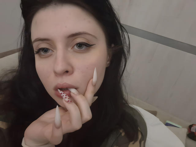 Close-up of DanielleZiego, pale-skinned with dark hair, blue eyes, and long white nails, sucking on a red and white patterned lollipop, looking directly at the camera with a slightly open mouth.