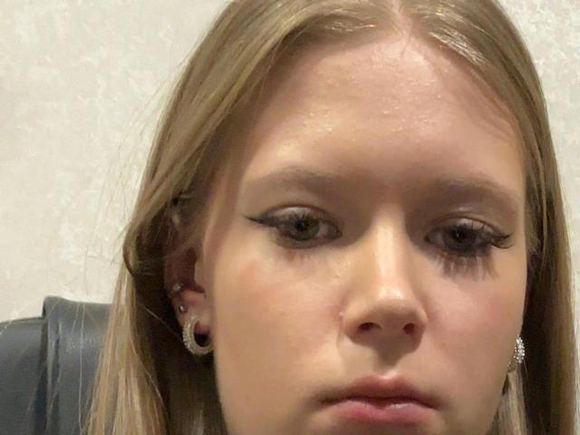 LillianaWhang's face, showing blonde hair, green eyes with thick eyelashes, wearing diamond earrings, and looking slightly bored or serious with a slight pinkish nose.