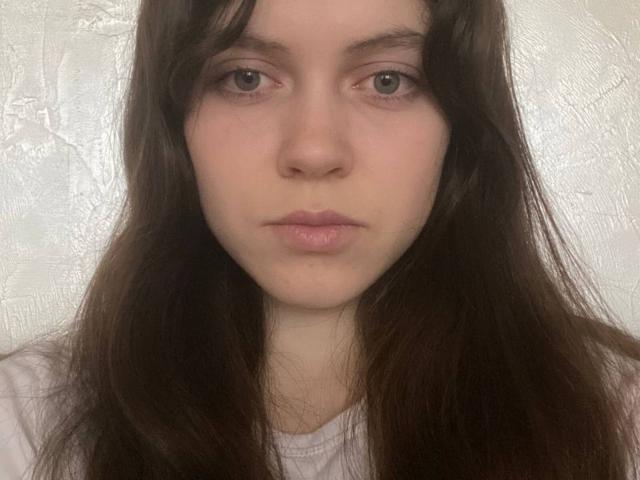 BellPrisco has long brown hair, light skin, and a neutral expression with slightly puffed pink lips. She's wearing a white shirt, looking directly at the camera.