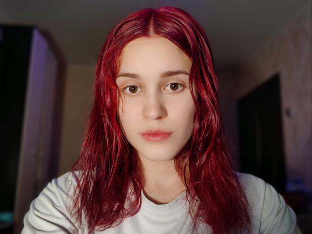 Cheri Worstell tiene el cabello rojo con un brillo que simula estar mojado, piel pálida, ojos marrones y una expresión neutra. Lleva una camisa blanca, con una ligera sombra en su lado derecho..