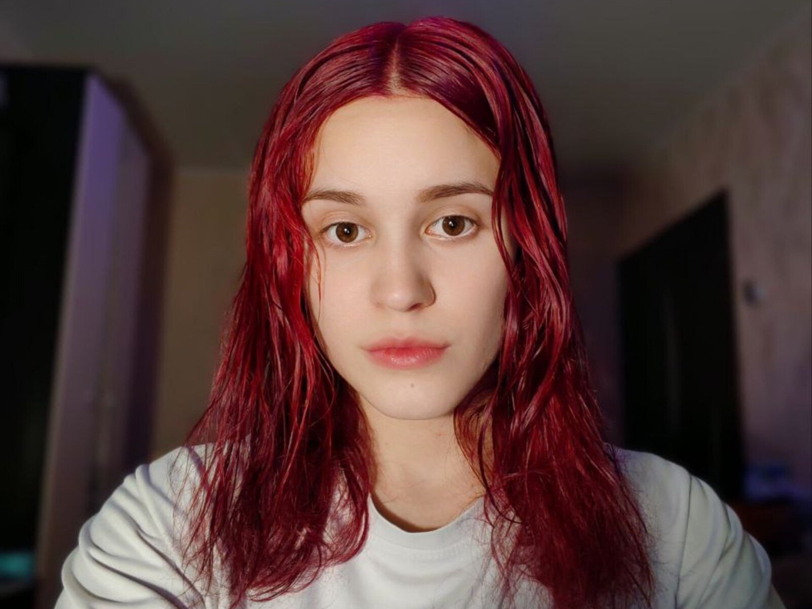 CheriWorstell tem cabelo vermelho com aparência molhada, pele clara, olhos castanhos e uma expressão neutra. Ela está vestindo uma camisa branca, com uma leve sombra no lado direito dela..