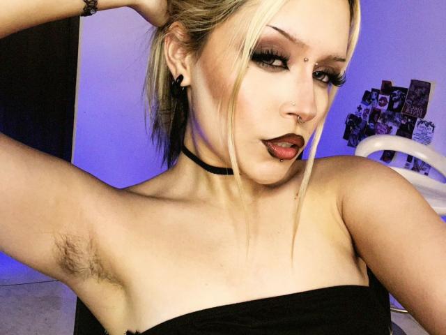 Fotografia ravvicinata di ScorpioGoddess, una donna bionda con rossetto rosso, un collare nero e ascelle scoperte, che guarda. con un'espressione seducente rivolta alla telecamera, alzando il braccio destro, sfondo illuminato di blu.