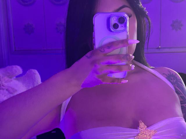 Primer plano de KatianPierce tomando una selfie con un teléfono inteligente, con iluminación azul-morada. KatianPierce tiene el pelo largo y negro, lleva un sujetador blanco, tiene un tatuaje en el hombro, uñas moradas y se puede ver el pene. Calcomanía de estrella brillante.