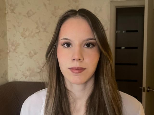 Fotografia em close-up de AugustineKerzer, com cabelo castanho liso, pele clara e olhos castanhos. Ela está vestindo uma blusa branca, olhando diretamente para a câmera com uma expressão neutra. O fundo apresenta um papel de parede floral e uma porta escura..