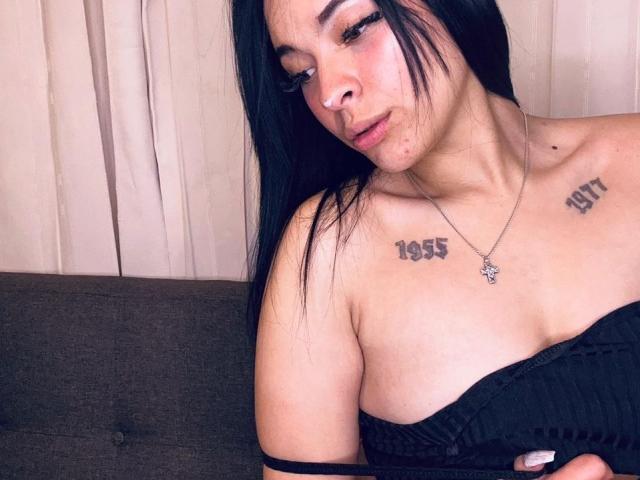 Photographie de MiaMillerr69, avec de longs cheveux noirs, regardant vers le bas avec une expression séduisante. Elle a des tatouages sur les épaules, un haut noir sans bretelles et un collier. La tension sexuelle est forte..