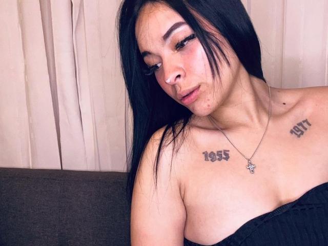 Fotografie von MiaMillerr69, einer attraktiven Frau mit langen, schwarzen Haaren, heller Haut und Tattoos an Schulter und Brust. Sie trägt ein schwarzes Top ohne Träger und blickt mit einem verführerischen Ausdruck nach unten..