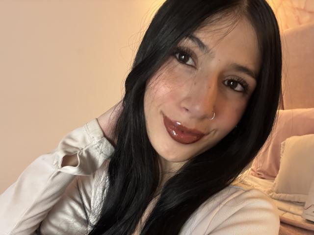 Close-up de Sophie Beaumont, uma jovem com cabelos longos e pretos, olhos castanhos e lábios vermelhos brilhantes, vestindo uma camisa branca. Sorrindo, com um piercing no nariz, sentada em uma cama. Tensão sexual.