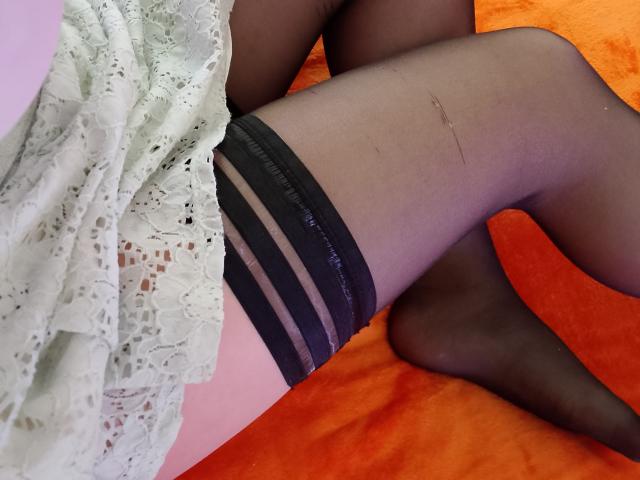 Close-up of Marriam's lower body, showing her legs crossed on an orange surface. She's wearing black lace-top stockings with a white, lace-trimmed dress. A visible scar runs down her thigh.