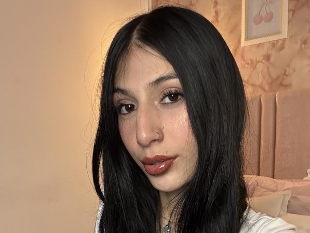 Nahaufnahme von Sophie Beaumont, einer jungen Frau mit langen, schwarzen Haaren, heller Haut und dunklen Augen, die ein weißes Hemd trägt. Nasenring und glänzliche Lippen, sie blickt verführerisch in die Kamera..