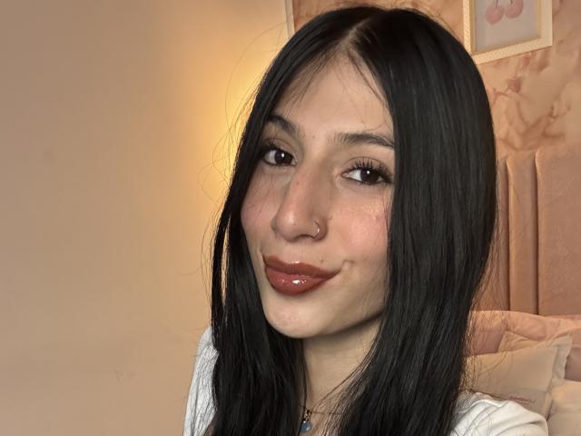 Fotografia di Sophie Beaumont, una giovane donna con lunghi capelli neri, pelle chiara e labbra marroni lucide, che sorride leggermente. con un piccolo piercing al naso, che indossa una maglietta bianca, in una camera da letto con una luce soffusa..