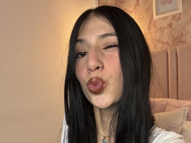 Fotografia de Sophie Beaumont com cabelos longos e pretos, piscando e franzindo seus lábios vermelhos e brilhantes. Ela tem um piercing no nariz e veste uma blusa branca. O fundo é um quarto com uma cabeceira estofada..